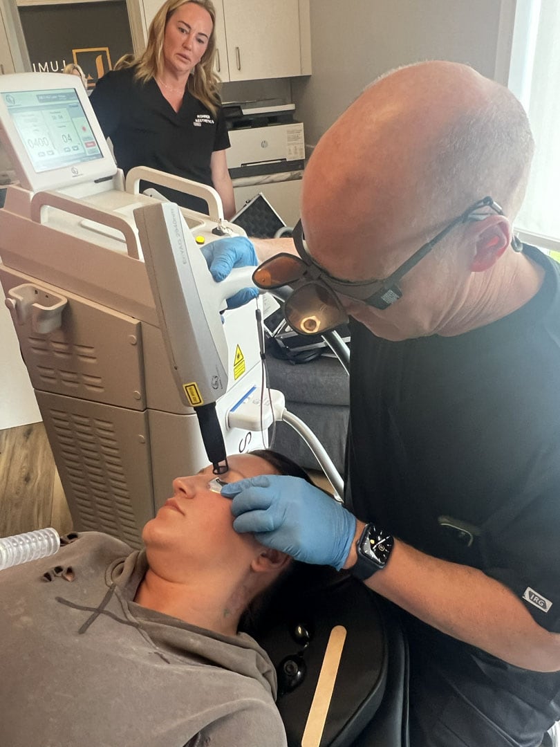 A person lies on a reclined chair receiving a facial laser treatment from a practitioner wearing blue gloves and protective eyewear. Another person in a black shirt observes in the background. Various medical equipment is visible around them.