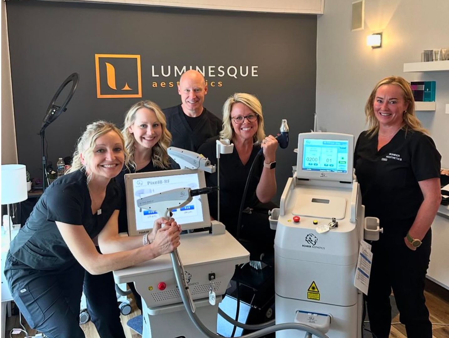 A group of five smiling people in black scrubs stand around aesthetic medical equipment, including laser devices, in a treatment room with the sign "Luminesque Aesthetics" on the wall. The environment is well-lit and professional.