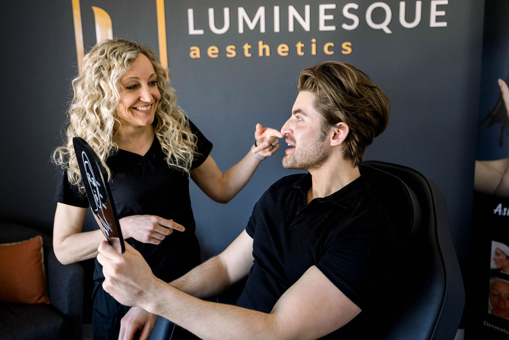 A woman with curly blonde hair, wearing a black outfit, smiles while pointing at the face of a seated man with light brown hair. The man, also dressed in black, holds a mirror, looking at his reflection. The background displays the text "LUMINESQUE aesthetics.