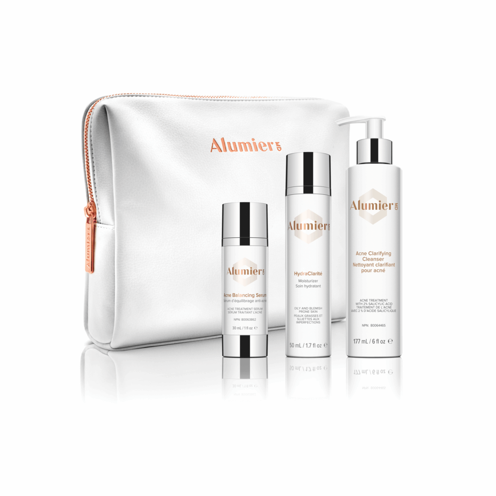 A white AlumierMD cosmetic bag with rose gold zipper is displayed with three AlumierMD skincare products in white bottles and silver accents arranged in front of it.